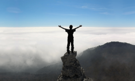 Person standing on the top of a rock stack on top of a mountain with outstretched arms
